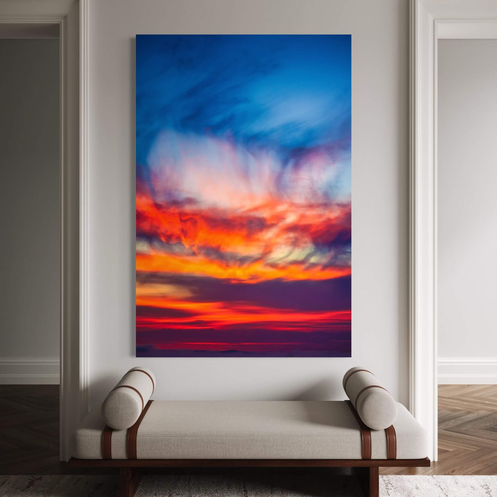 Smoldering Clouds 60 x 40 high-definition metal print by Abigail Diane Photography displayed as large statement wall art above an upholstered bench in an elegant contemporary interior, featuring sweeping cobalt blue, crimson red, fiery orange, violet, and luminous golden clouds at sunset over the Pacific Ocean in Southern California.