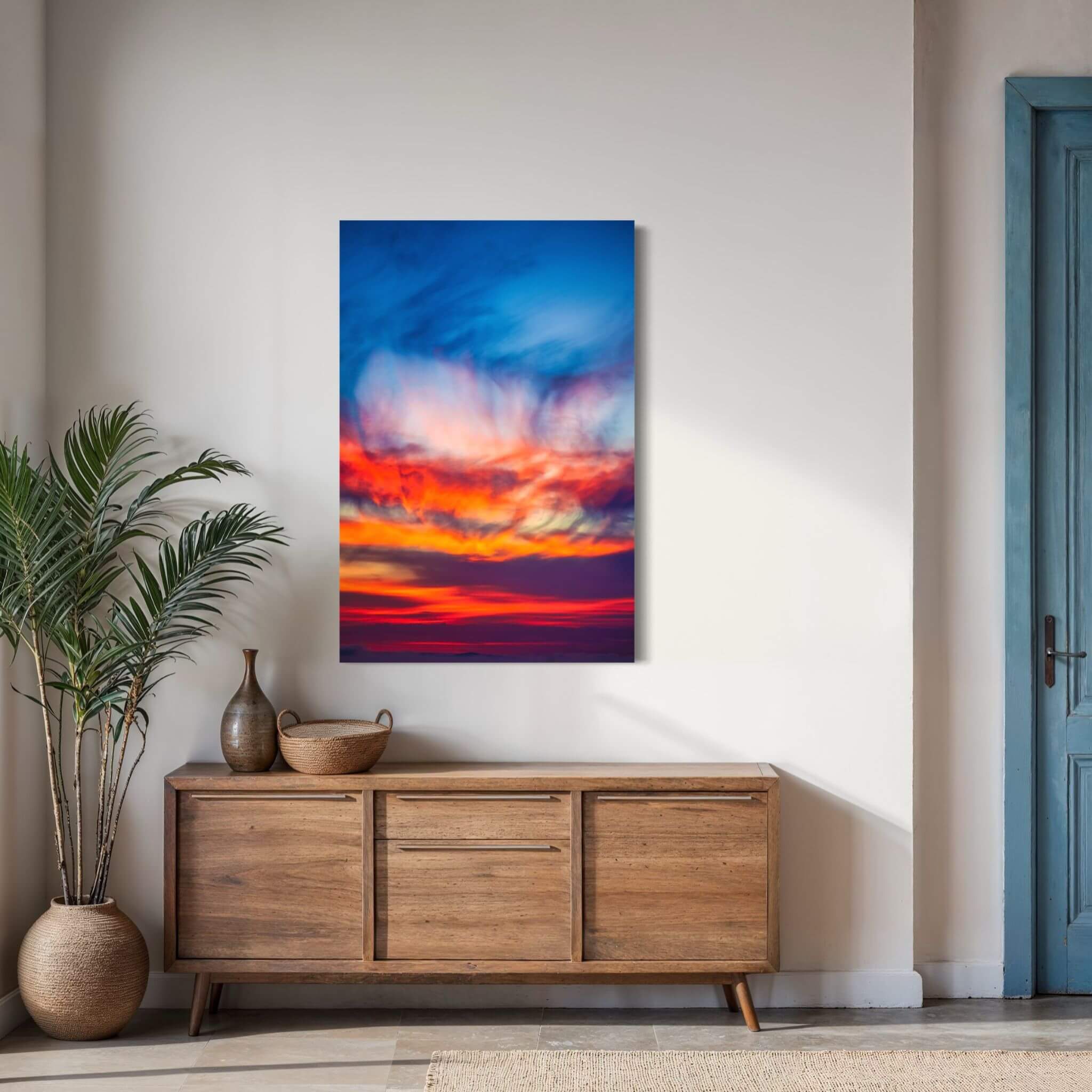 Smoldering Clouds high-definition metal print by Abigail Diane Photography displayed above a natural wood console in a bright contemporary entryway, featuring vivid cobalt blue, crimson red, fiery orange, purple, and golden sunset clouds over the Pacific Ocean in Southern California.