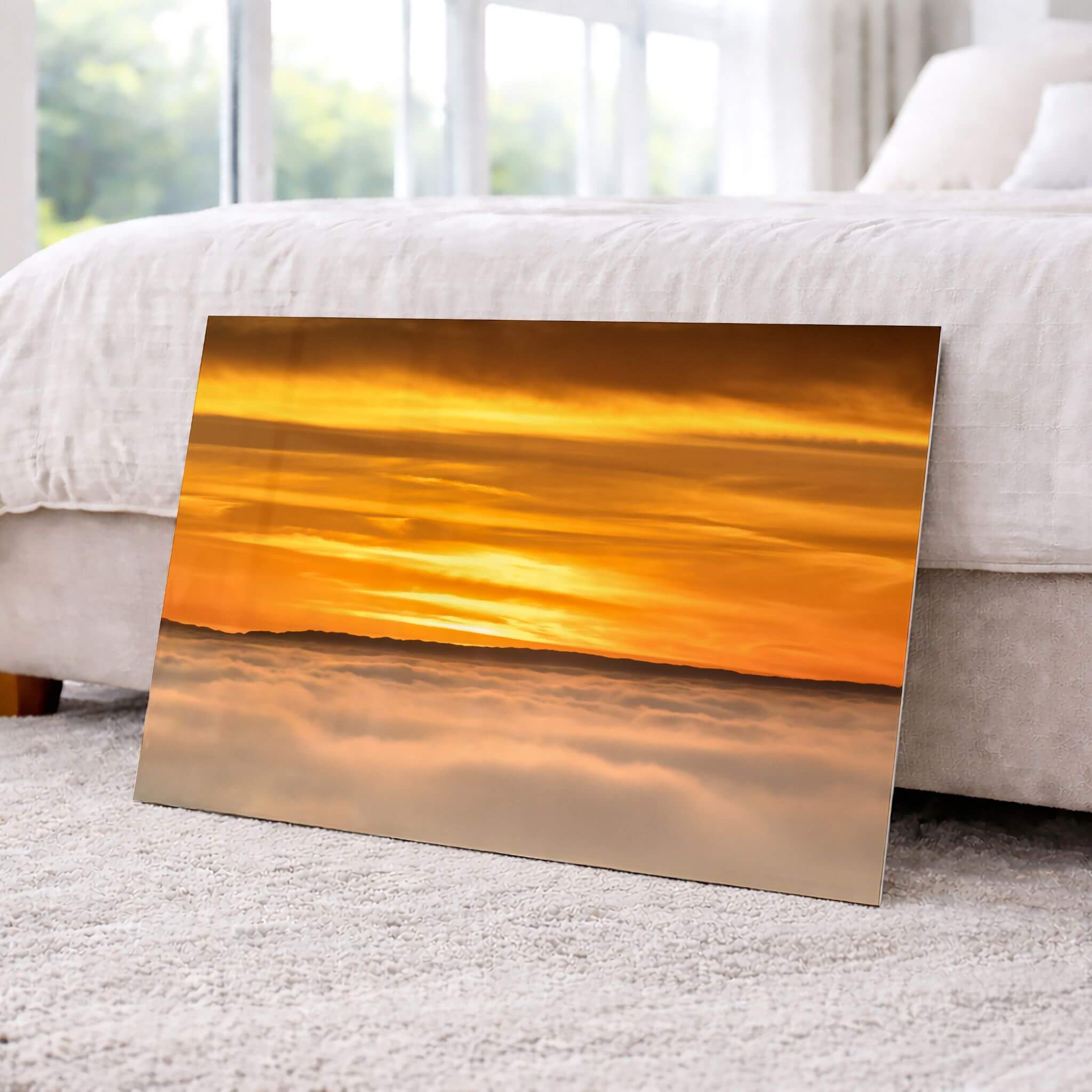 HD metal print of Soaring Above the Clouds by Abigail Diane Photography leaning against a bed in a bright bedroom, highlighting a radiant golden sunset above soft cloud layers with exceptional color and detail