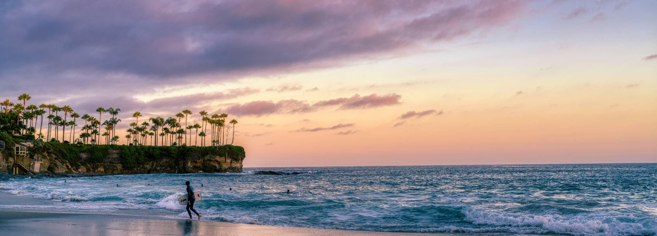 Surfing a California Sunset  print featuring a lone surfer walking along Crescent Beach in Laguna Beach, California beneath soft pink sunset clouds with gentle Pacific Ocean waves and reflective shoreline. Premium wall art by Abigail Diane Photography.