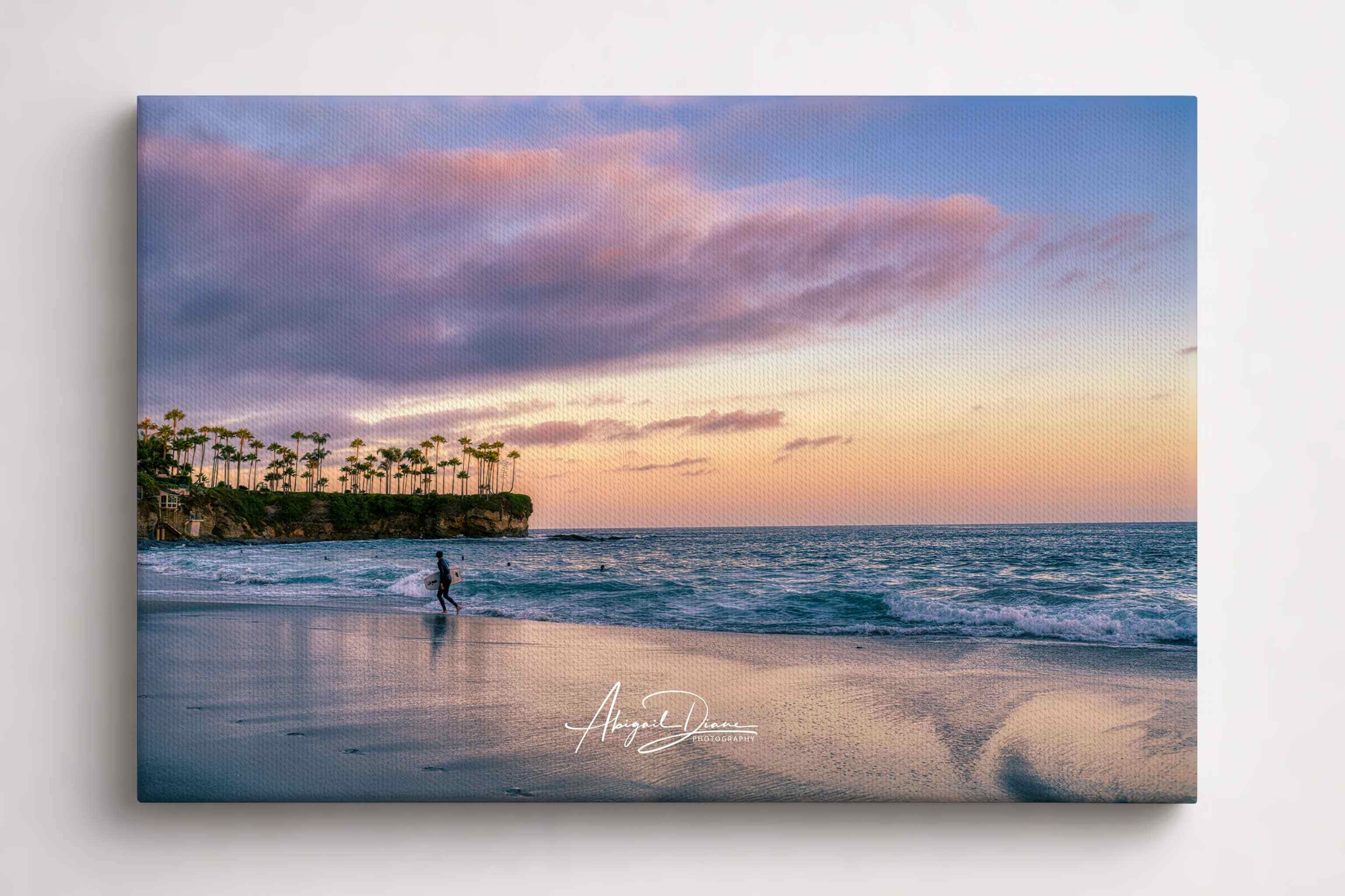 Surfing a California Sunset canvas print featuring a lone surfer walking along Crescent Beach in Laguna Beach, California beneath soft pink sunset clouds with gentle Pacific Ocean waves and reflective shoreline. Gallery wrapped canvas wall art by Abigail Diane Photography.