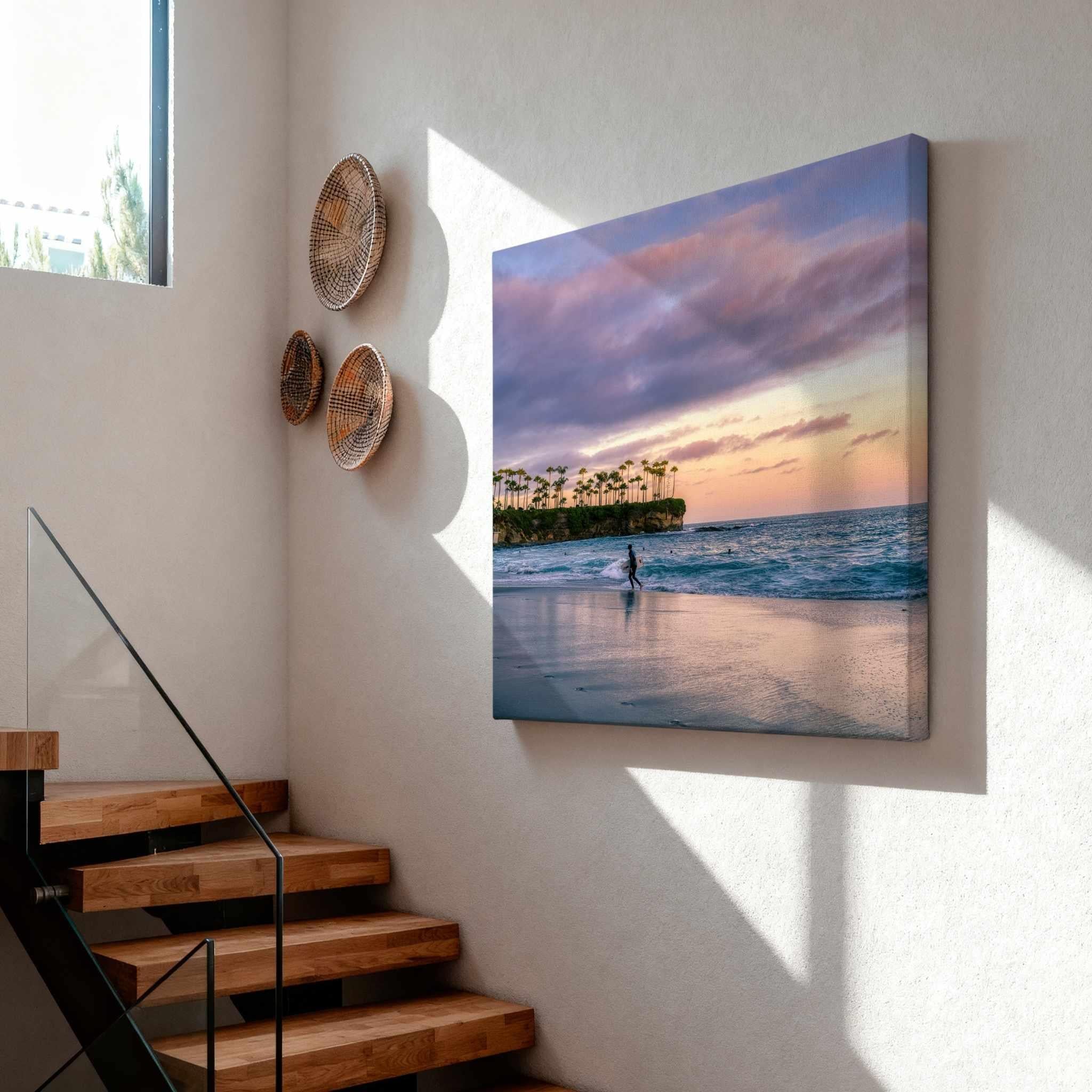 Surfing a California Sunset gallery-wrapped canvas print displayed on a modern staircase wall, featuring a lone surfer at Crescent Beach beneath pastel sunset skies in Laguna Beach, California.