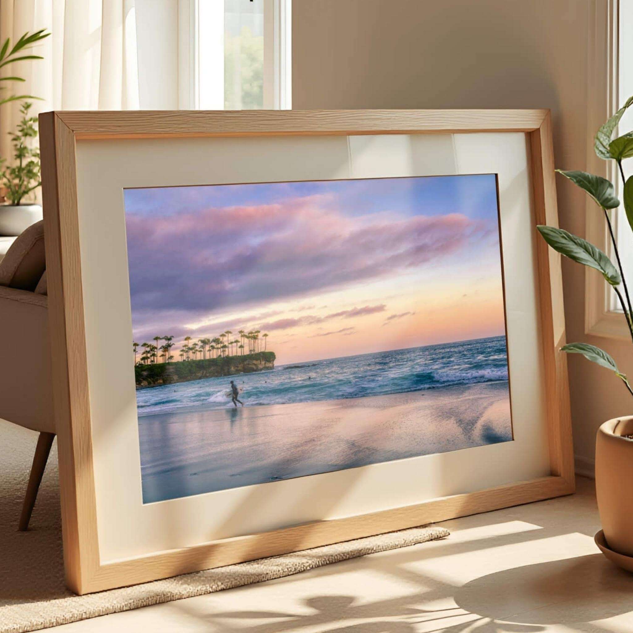 Framed fine art print of Surfing a California Sunset displayed in a natural oak frame with a white mat, featuring a lone surfer walking along the shoreline at Laguna Beach, California beneath soft pink and lavender sunset clouds. Gentle Pacific Ocean waves and reflective wet sand create a calming coastal wall art piece by Abigail Diane Photography, available in multiple sizes and premium framing options.