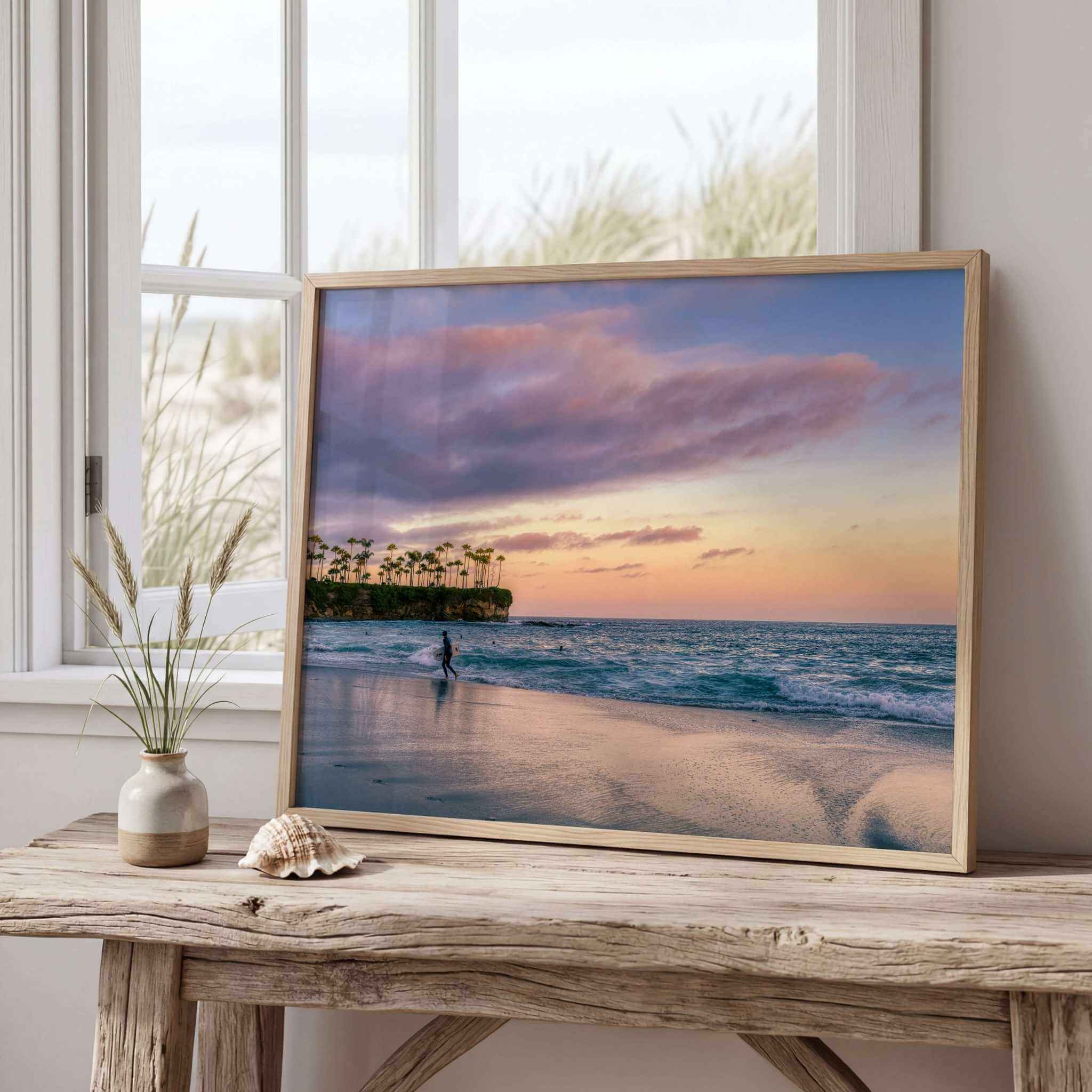 Framed Surfing a California Sunset fine art photography print displayed on a rustic wooden console beside a bright window in a coastal-inspired home. Captured at Crescent Beach, Laguna Beach, California, this peaceful Pacific Ocean scene features a lone surfer, gentle waves, reflective wet sand and soft pink and lavender sunset clouds, creating calming coastal wall art. Available as fine art prints, framed prints and canvas in multiple sizes by Abigail Diane Photography.