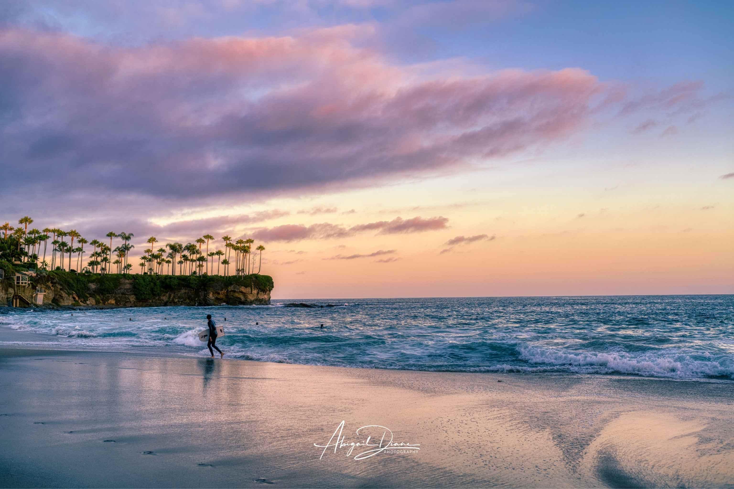 Surfing a California Sunset fine art photography print featuring a lone surfer walking along the shoreline at Laguna Beach, California beneath soft pink and lavender sunset clouds. Gentle Pacific Ocean waves, reflective wet sand and the iconic palm-lined cliffs create a peaceful coastal landscape available as fine art prints, framed wall art and canvas in multiple sizes.