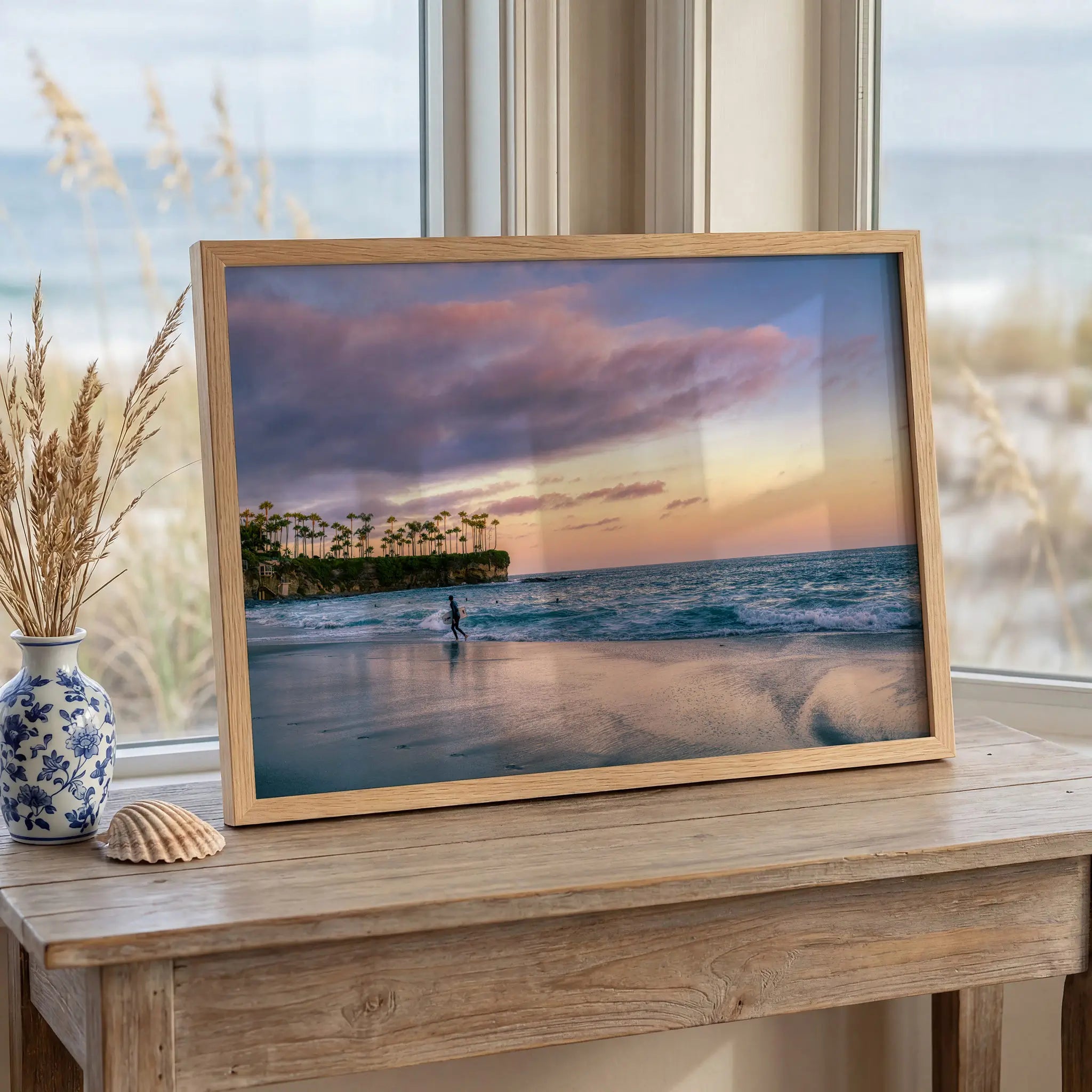 Surfing a California Sunset framed print featuring a lone surfer along the shoreline beneath a soft pink and golden sunset in Laguna Beach, California.