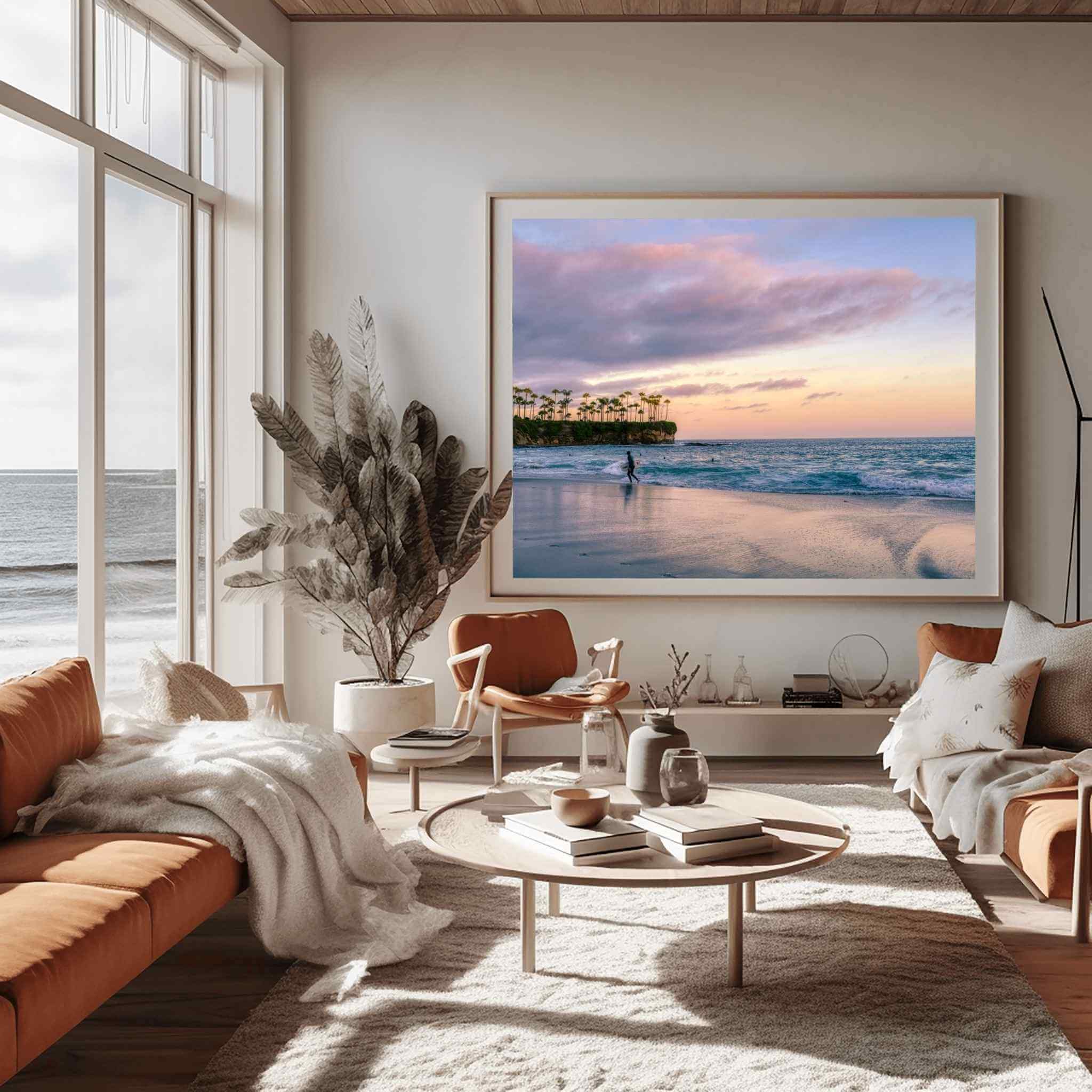 Large framed Surfing a California Sunset fine art photography print displayed in a warm contemporary coastal living room overlooking the ocean. Captured at Laguna Beach, California, this peaceful wall art features a lone surfer walking along the shoreline beneath soft pink and lavender sunset clouds, with gentle Pacific Ocean waves, reflective wet sand and the iconic palm-lined cliffs creating a serene coastal atmosphere. Available as fine art prints, framed wall art and canvas in multiple sizes by Abigail 